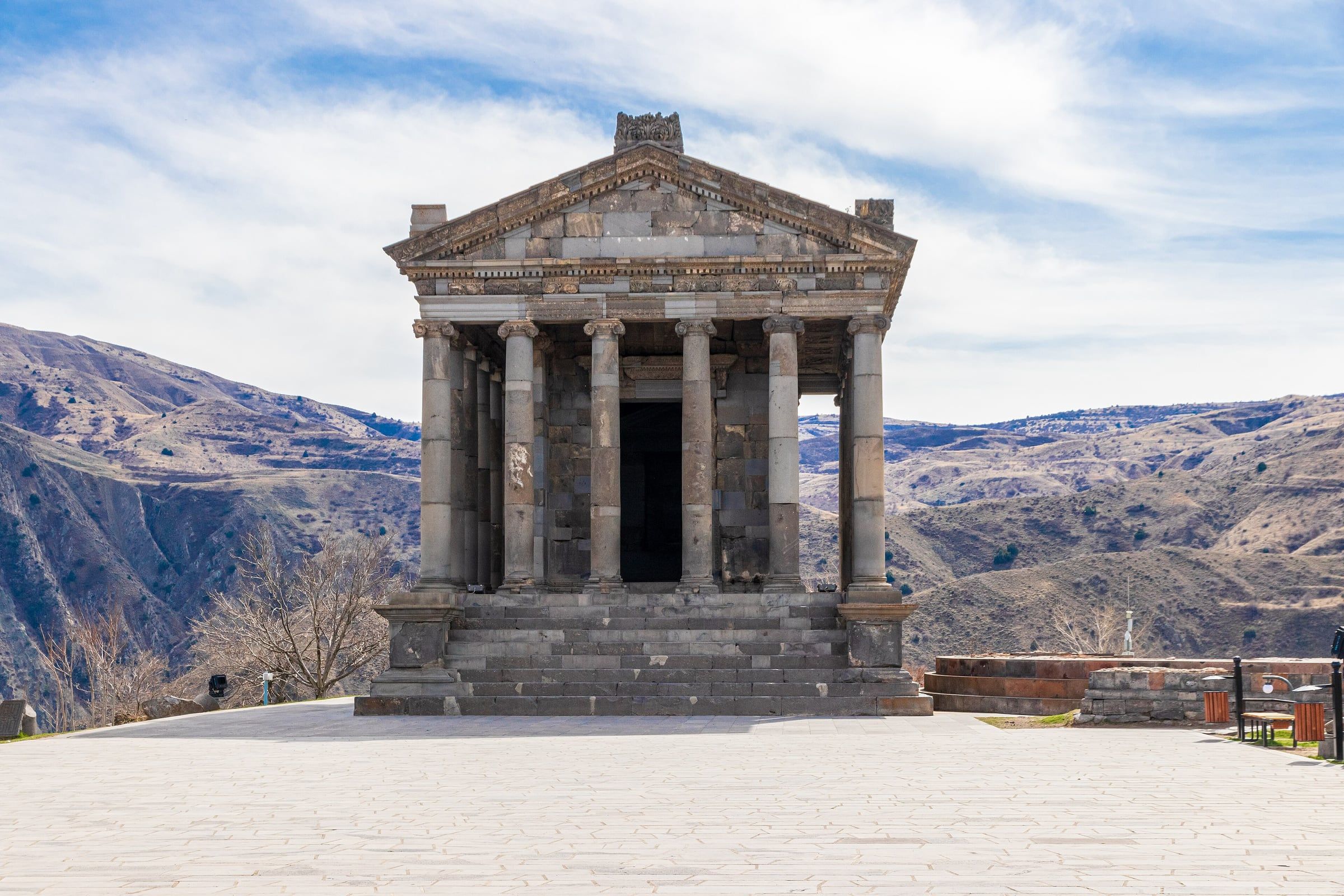 The colonnaded stone temple stands on a paved platform, with dry hills and ridges rising behind it under a cloudy sky.