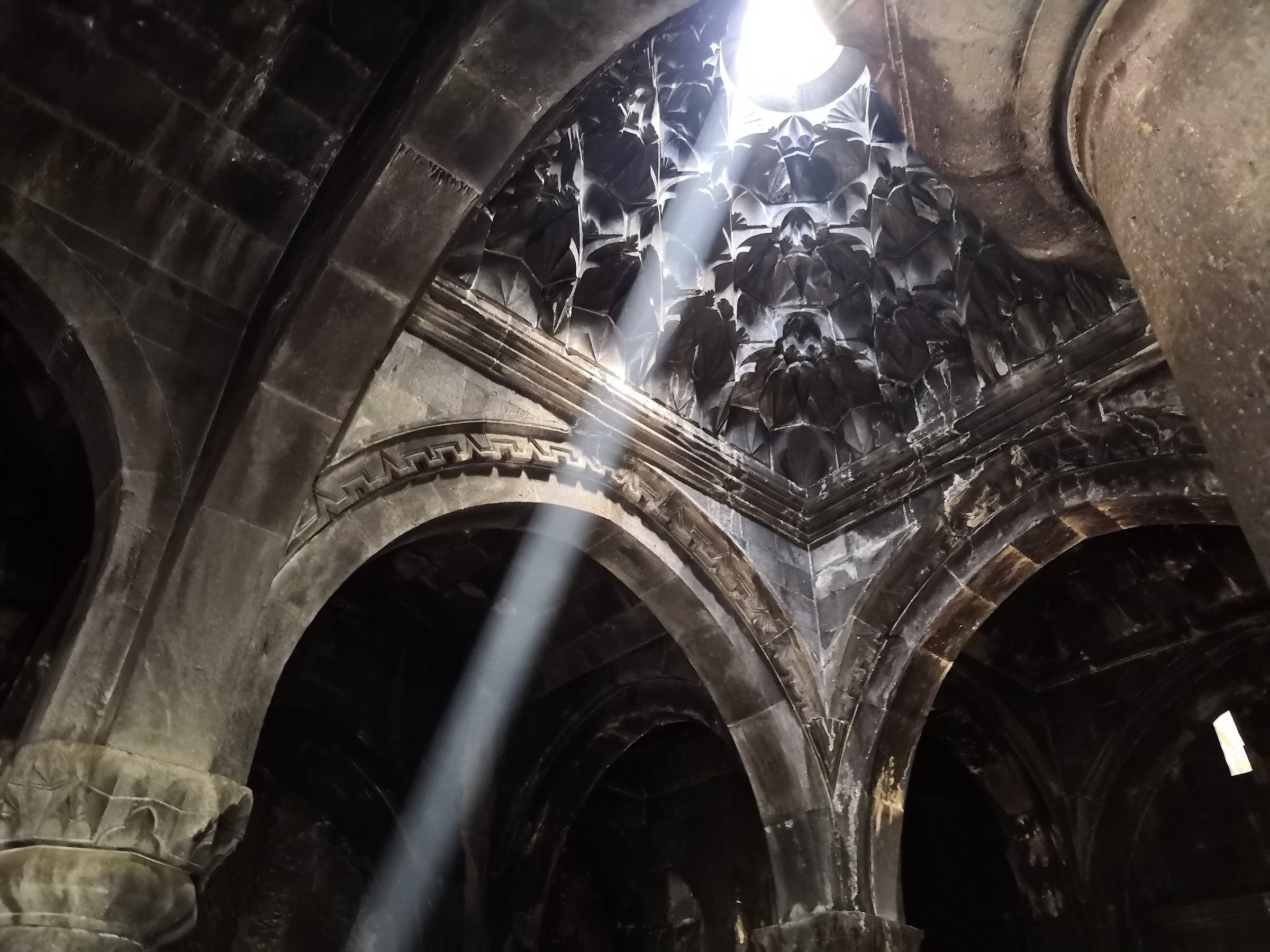 Light falls through an opening in a carved stone dome, lighting the ribbed arches of a vaulted hall.