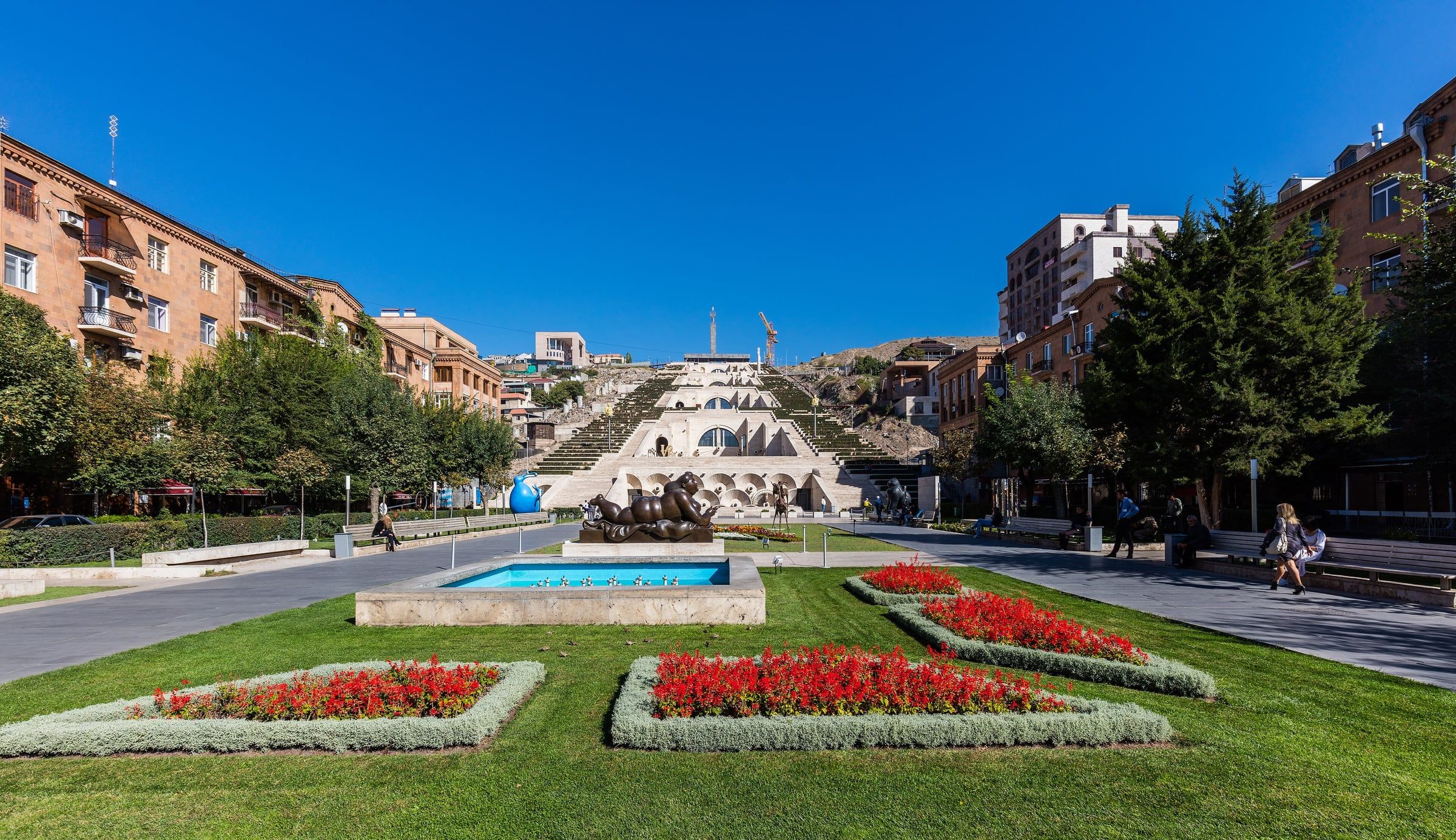 Green lawns, red flowerbeds and a fountain lead towards a wide stone staircase under a clear blue sky.